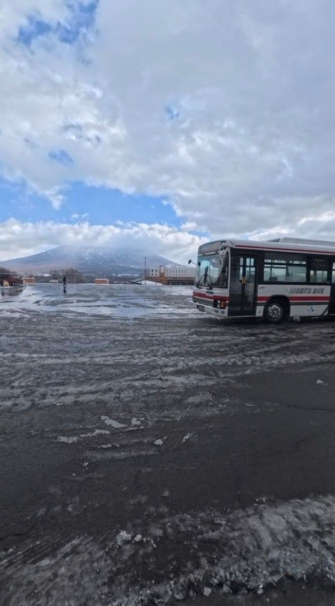 一輛 NISEKO BUS 停在融雪的停車場,後方是雲帶環繞的羊蹄山