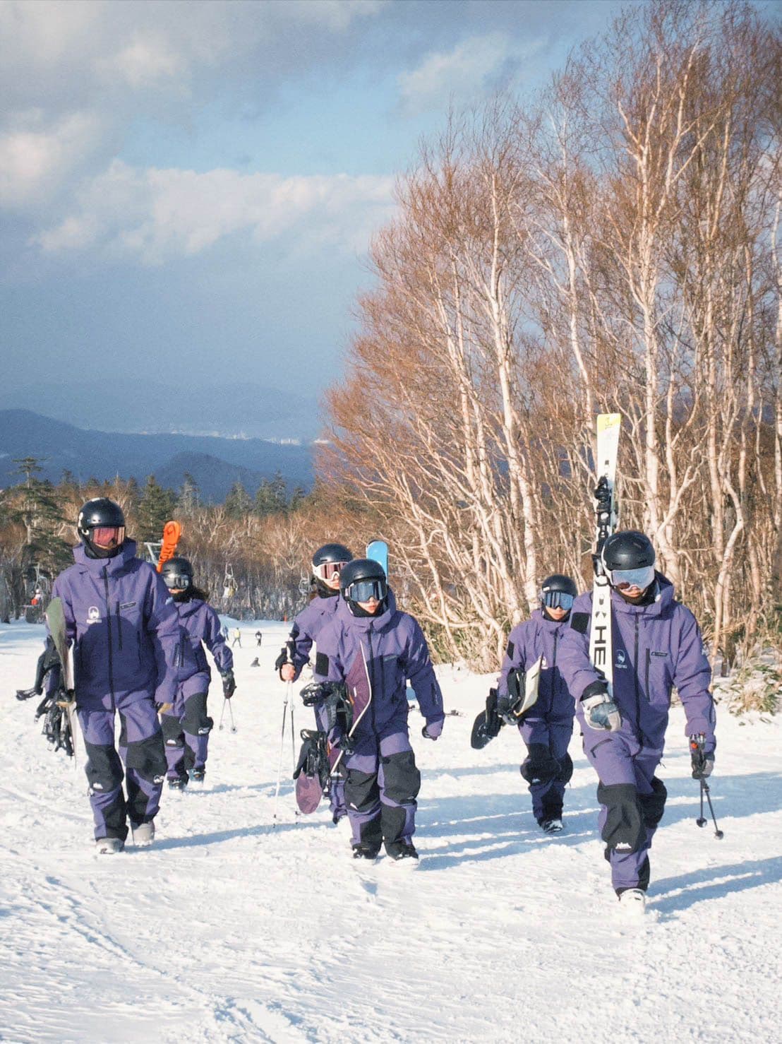 六位穿紫色 FUYU 制服的教練扛著雪具走在雪道上,背後是白樺林與遠山
