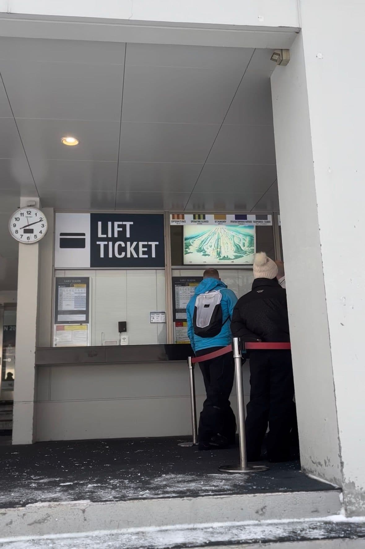 雪場的 LIFT TICKET 售票窗口,兩位客人正在看票價表,旁邊掛著纜車運行狀況看板